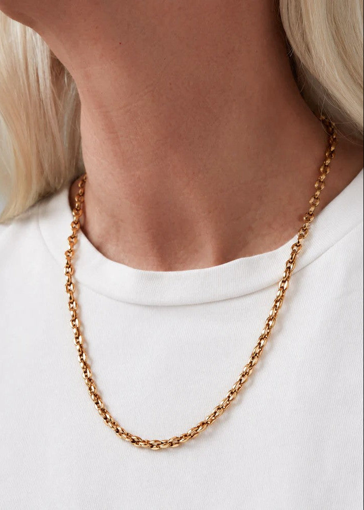 A close-up of a model's neck and upper chest wearing a delicate gold chain necklace over a white crew-neck top.