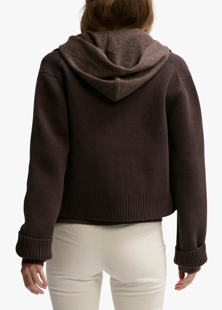 A person shown from the back wearing a dark brown ribbed knit hooded sweater with cuffed sleeves and cream-colored pants, against a plain light background.