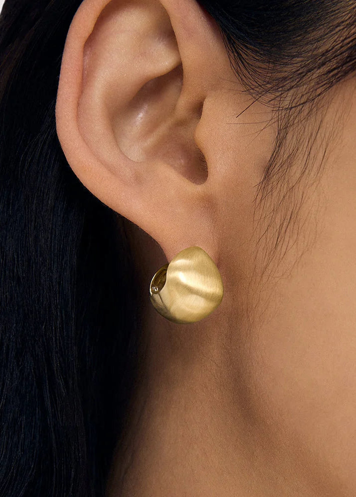 A close-up of a person's ear wearing a small, matte gold dome-shaped stud earring.
