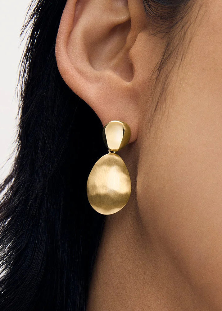 A close-up of a person's ear wearing a textured, organic-shaped gold drop earring against a light gray background.
