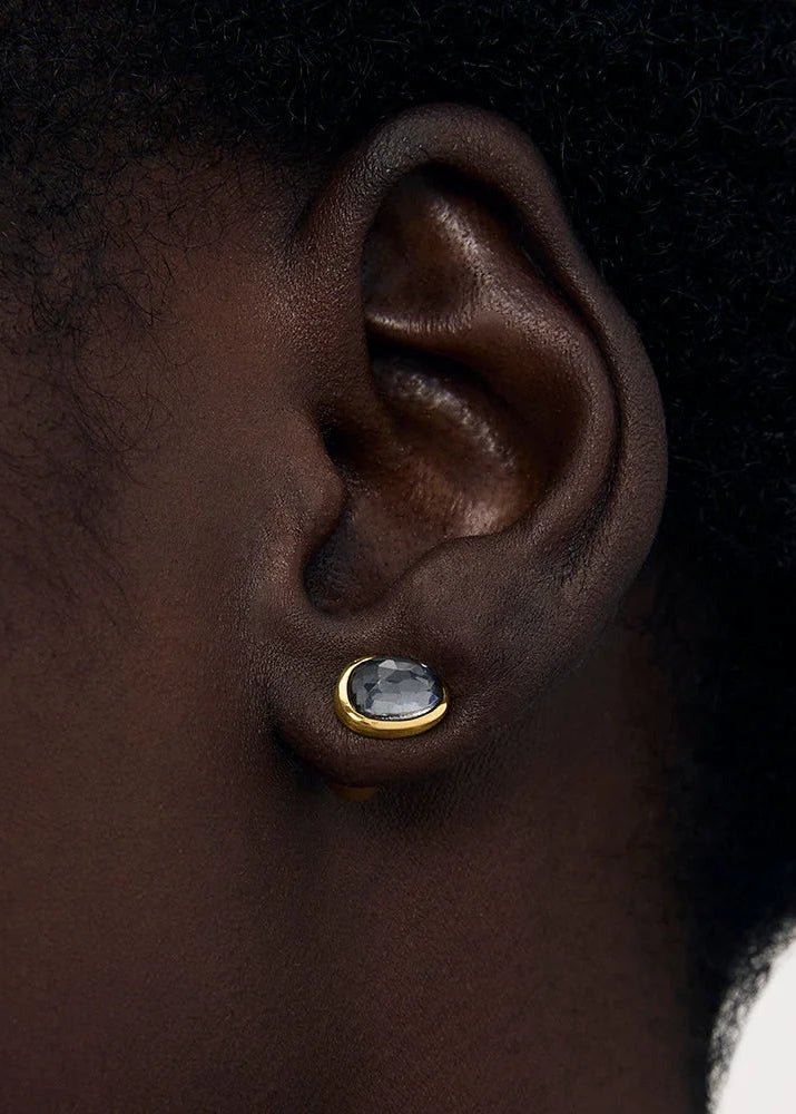A close-up of a person's ear wearing a small gold stud earring with a round clear gemstone.