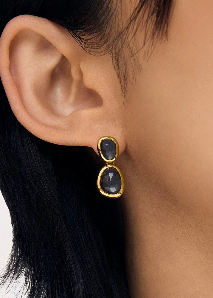 A close-up of a person's ear wearing a gold drop earring with two oval dark gray gemstones.