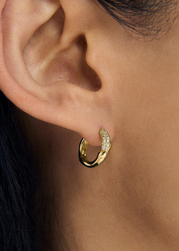 A close-up of a person's ear wearing a small gold huggie hoop earring with a row of clear pavé stones along the front curve.