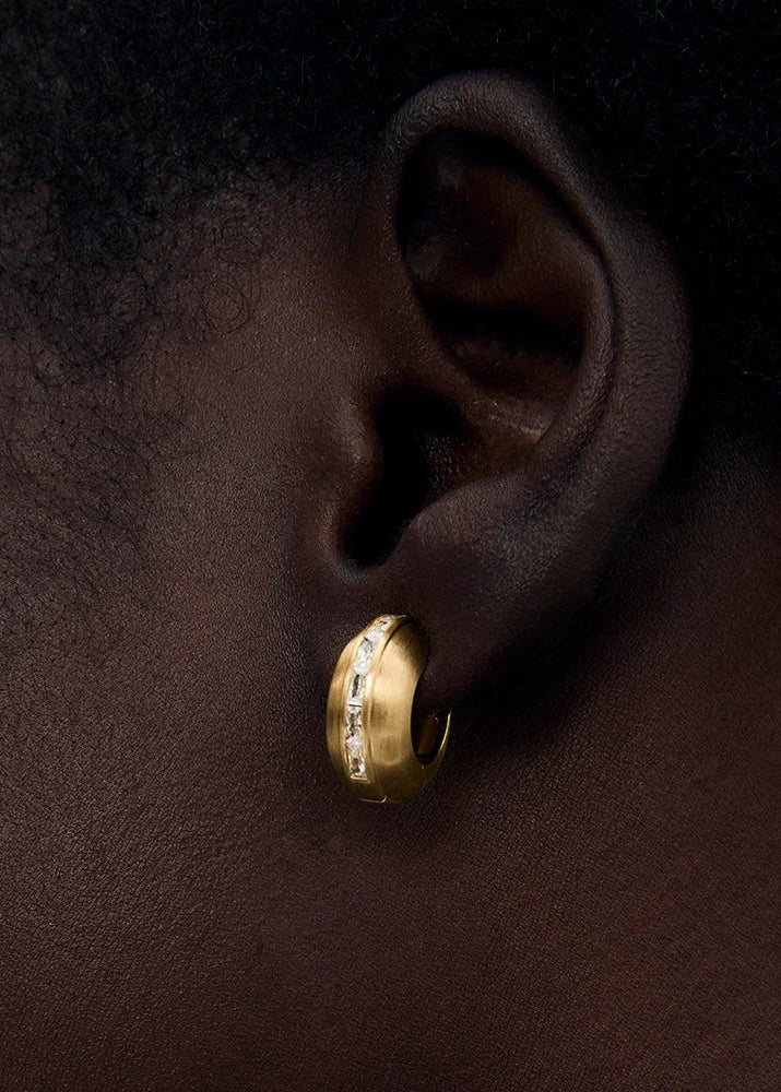 A close-up of a person's ear wearing a small gold huggie hoop earring with a row of clear stones along the front.