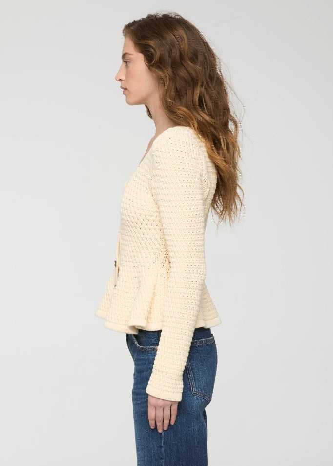 A woman shown in profile wearing a cream-colored, textured knit cardigan with a peplum hem and blue jeans, set against a plain light gray studio background.