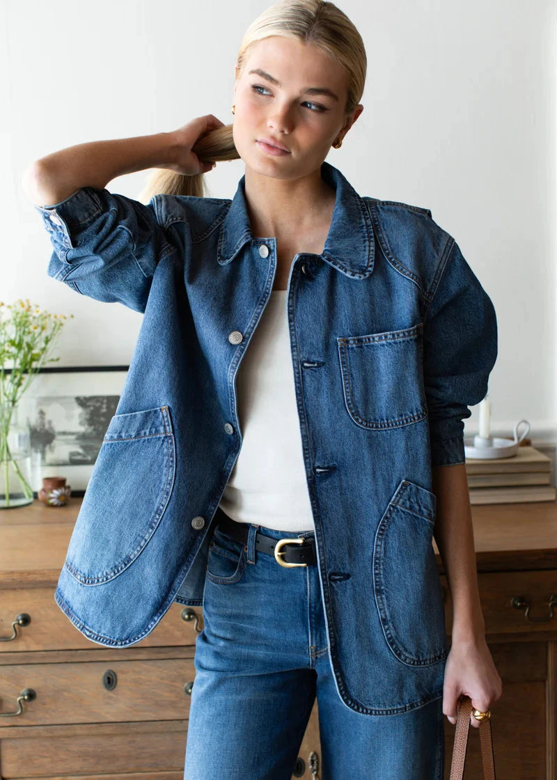 Woman wearing a denim jacket and jeans in a room with wooden furniture.