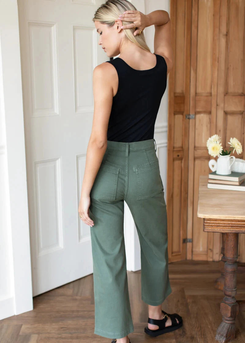Woman wearing a black sleeveless top and green pants standing in a room with wooden flooring and a table.
