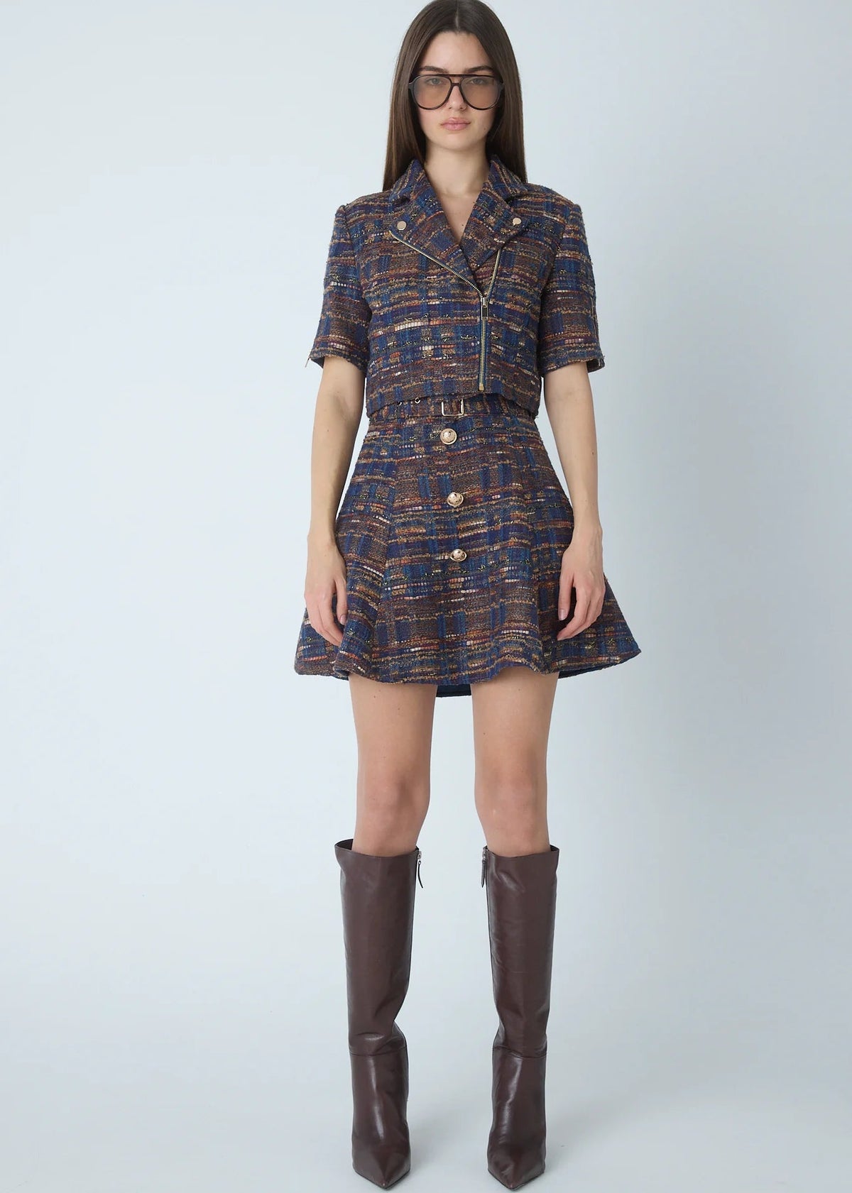 A model wearing a short-sleeve tweed mini dress with a shawl collar, front zipper, and belt, paired with dark brown knee-high boots against a light gray studio background.