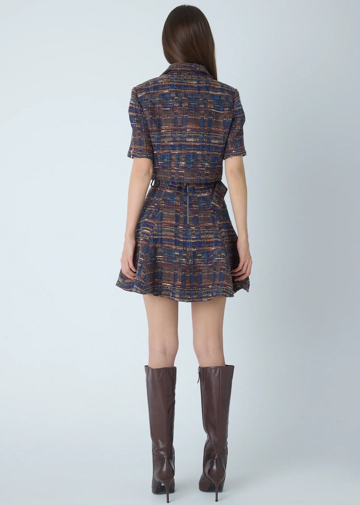 A model viewed from the back wearing a short-sleeve mini dress with a brown and blue plaid tweed pattern and matching tie belt, paired with brown knee-high boots against a light gray studio background.