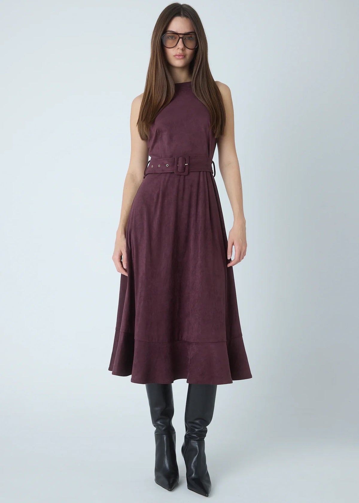 A woman modeling a sleeveless high-neck midi dress in a deep plum purple color, featuring a matching belt at the waist. She is also wearing black knee-high leather boots and glasses, standing against a light gray studio background.