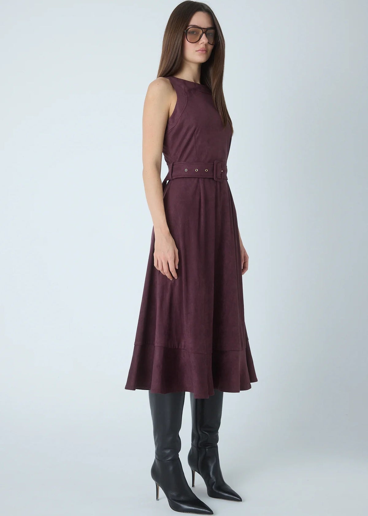 A woman modeling a sleeveless, high-neck, A-line midi dress in a deep plum purple color, styled with a matching belt and black pointed-toe heeled boots against a light gray studio background.