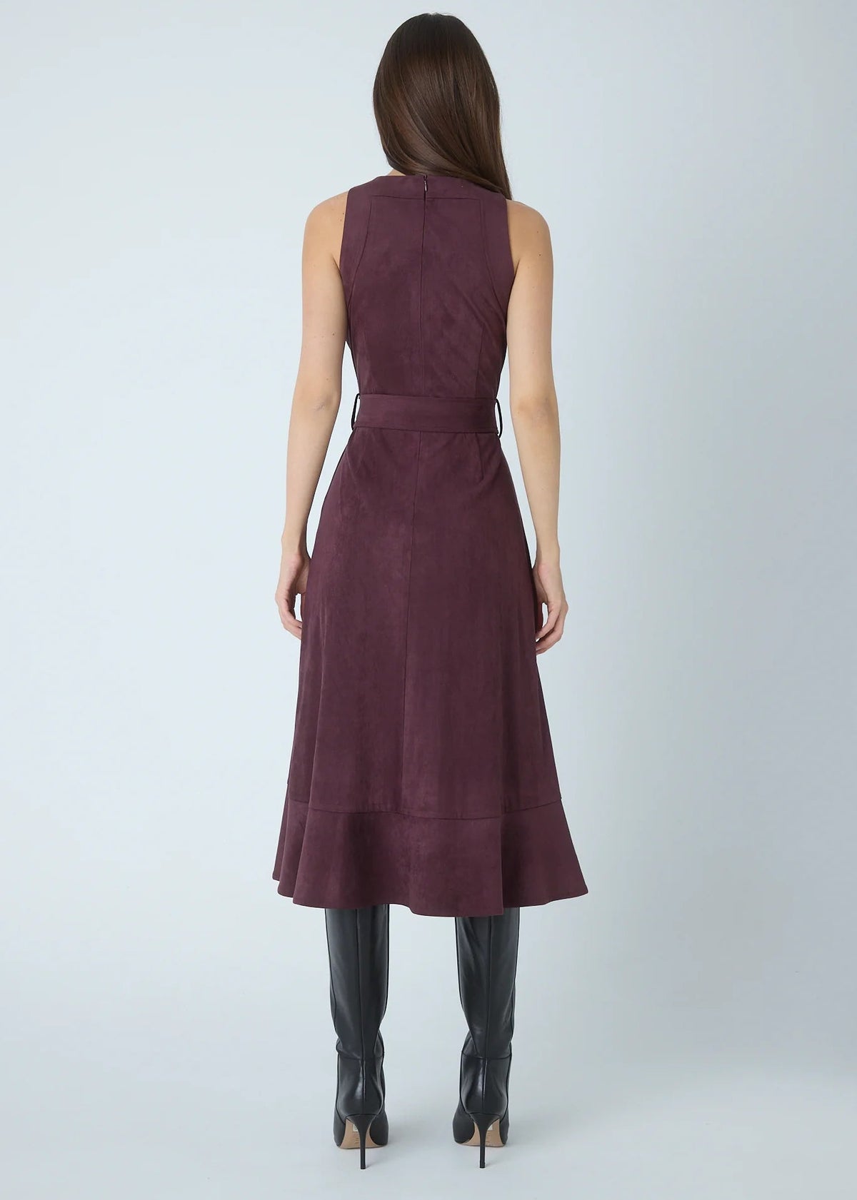 A woman viewed from the back wearing a sleeveless, knee-length dark plum dress with a matching waist tie, paired with black knee-high boots, against a light gray studio background.