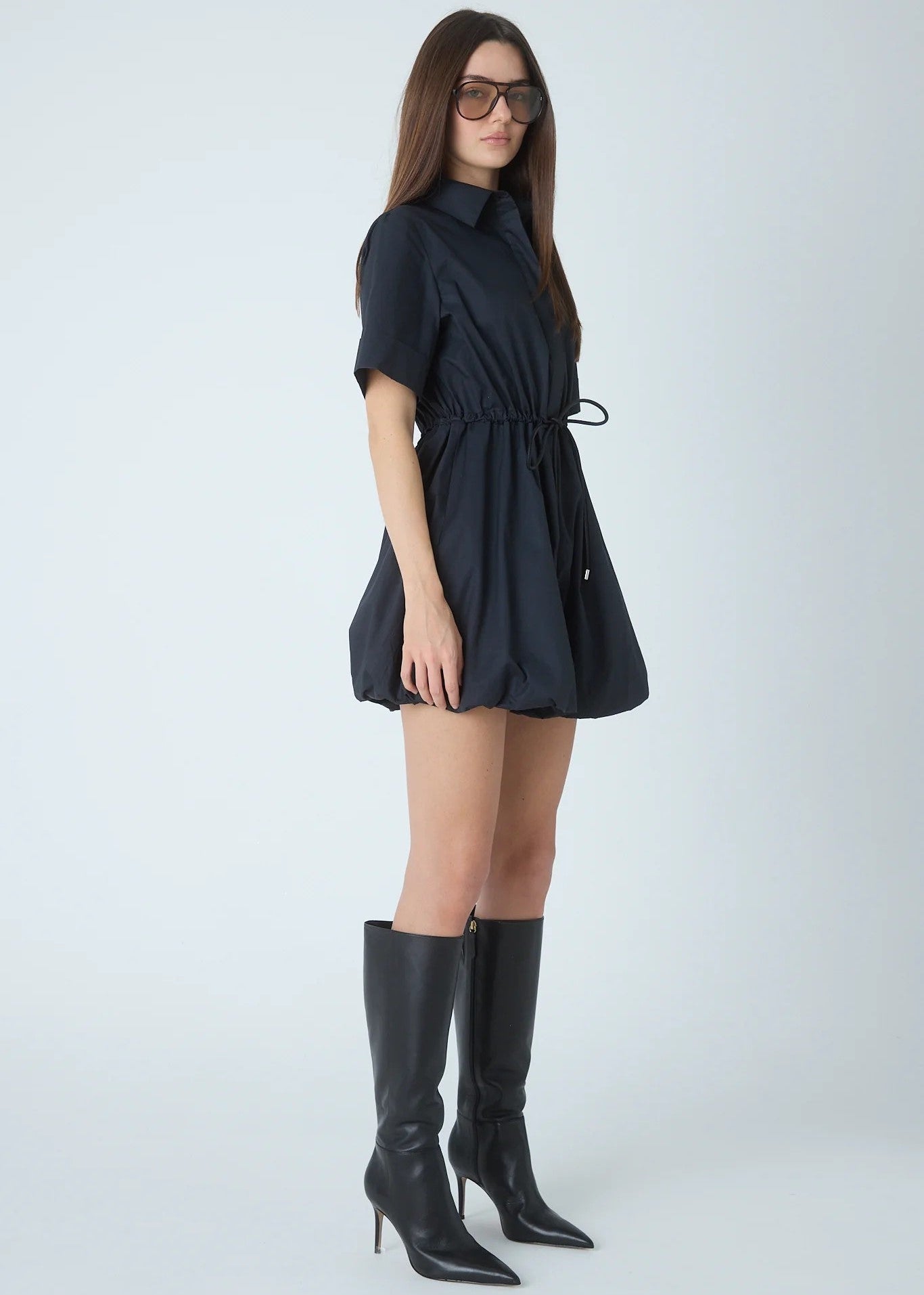 A woman modeling a short-sleeve black mini dress with a tie waist, paired with black knee-high heeled boots, against a light gray studio background.