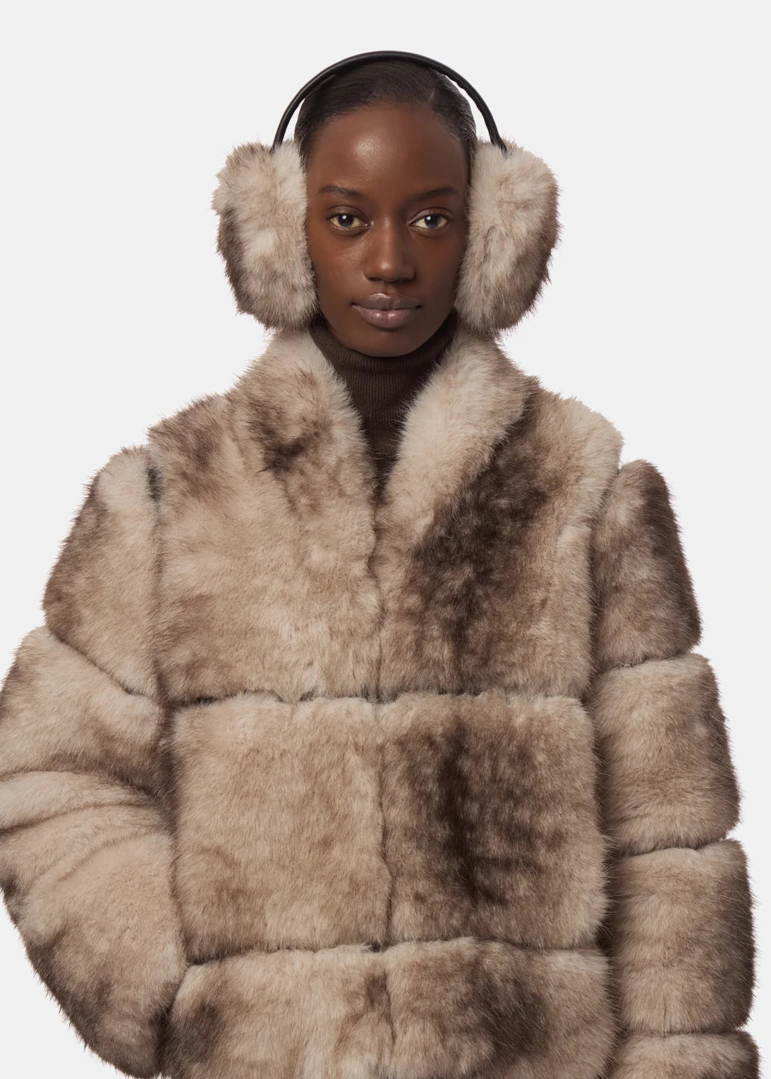 A model wearing a plush, patchwork-style faux fur coat in shades of beige and brown, paired with matching beige faux fur earmuffs with black earcups, against a solid light gray background.