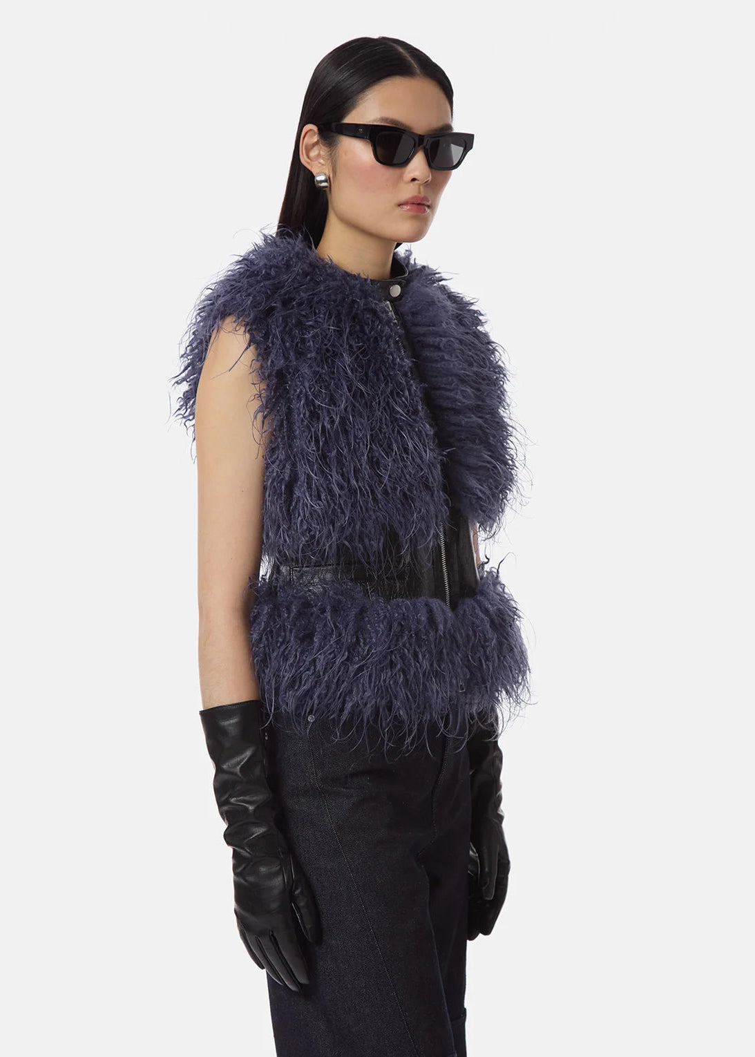 A model wearing a sleeveless, dark blue-gray shaggy faux-fur vest with a black belt, black leather gloves, and black sunglasses, set against a plain light gray background.