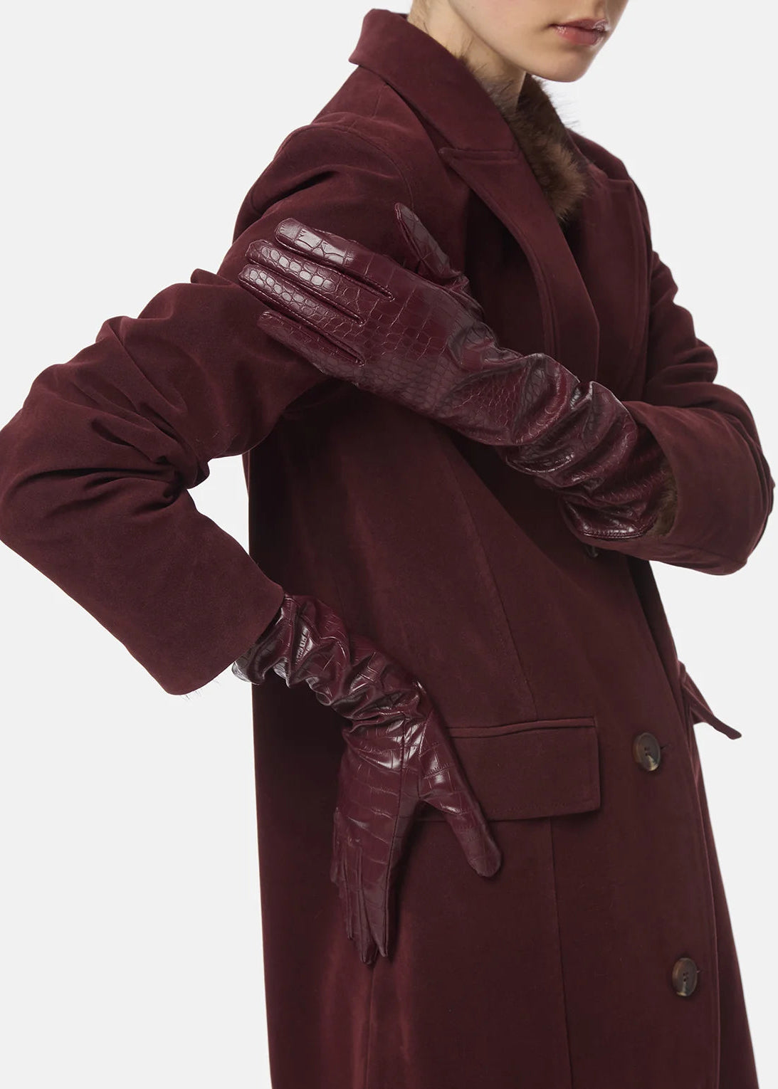 A model wearing a long burgundy wool-blend coat with dark burgundy leather gloves, shown from the chest down against a light gray studio background.