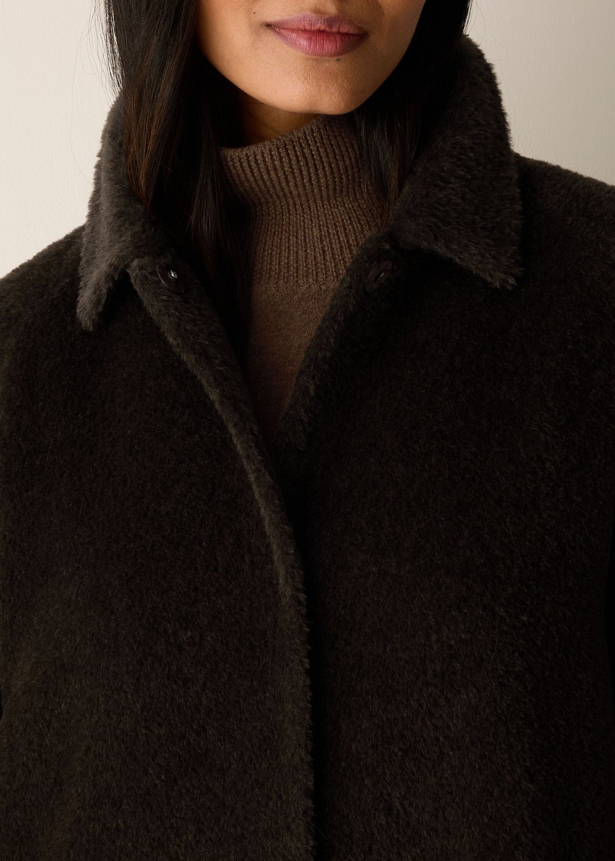 Close-up of a person wearing a dark brown fuzzy textured coat over a taupe ribbed turtleneck sweater, against a plain light background.