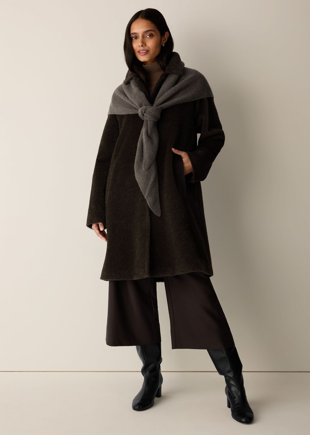 A woman models a dark brown wool coat with a contrasting dark gray shawl draped over her shoulders, paired with dark brown wide-leg trousers and black ankle boots, against a plain beige studio background.