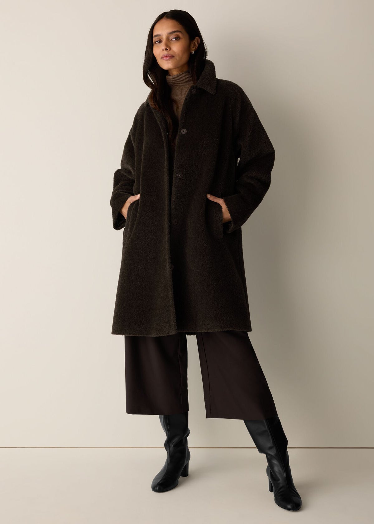 A woman models a dark brown textured wool-blend coat with a high collar and front buttons, styled with wide-leg dark trousers and black boots against a neutral beige studio background.