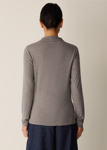 A woman viewed from the back wearing a heather gray long-sleeve mock neck top and dark blue pants, set against a plain light beige background.