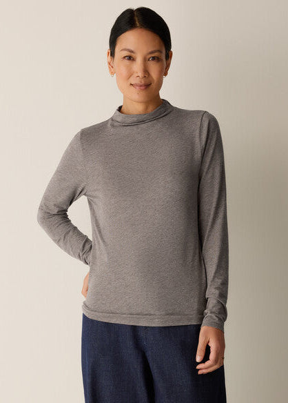 A woman modeling a long-sleeve gray mock neck top paired with dark blue pants, standing against a plain light beige background.