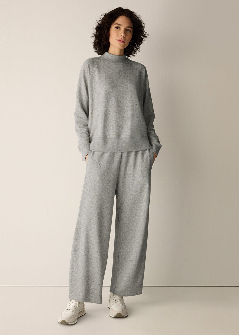 A woman modeling a light gray knit lounge set consisting of a mock-neck long-sleeve top and matching wide-leg pants, paired with white sneakers, against a neutral beige studio background.