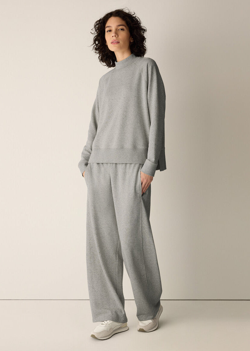 A woman modeling a light gray knit lounge set consisting of a long-sleeve mock-neck top and matching wide-leg pants, paired with white sneakers, against a plain beige studio background.