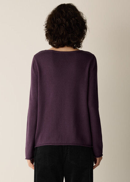 A person shown from the back wearing a long-sleeve knit top in a deep plum purple, paired with black pants, against a plain light beige studio background.