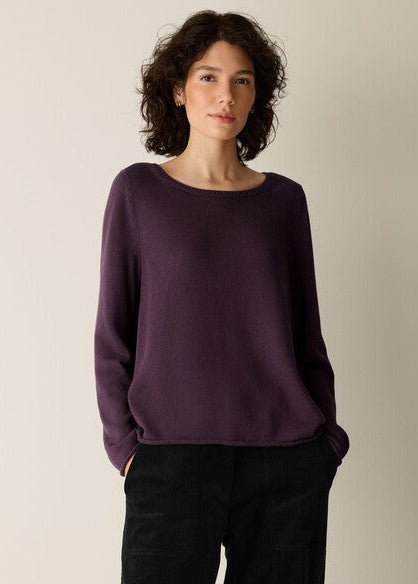 A woman with curly dark hair wearing a dark purple knit sweater and black pants, standing against a plain light beige studio background.