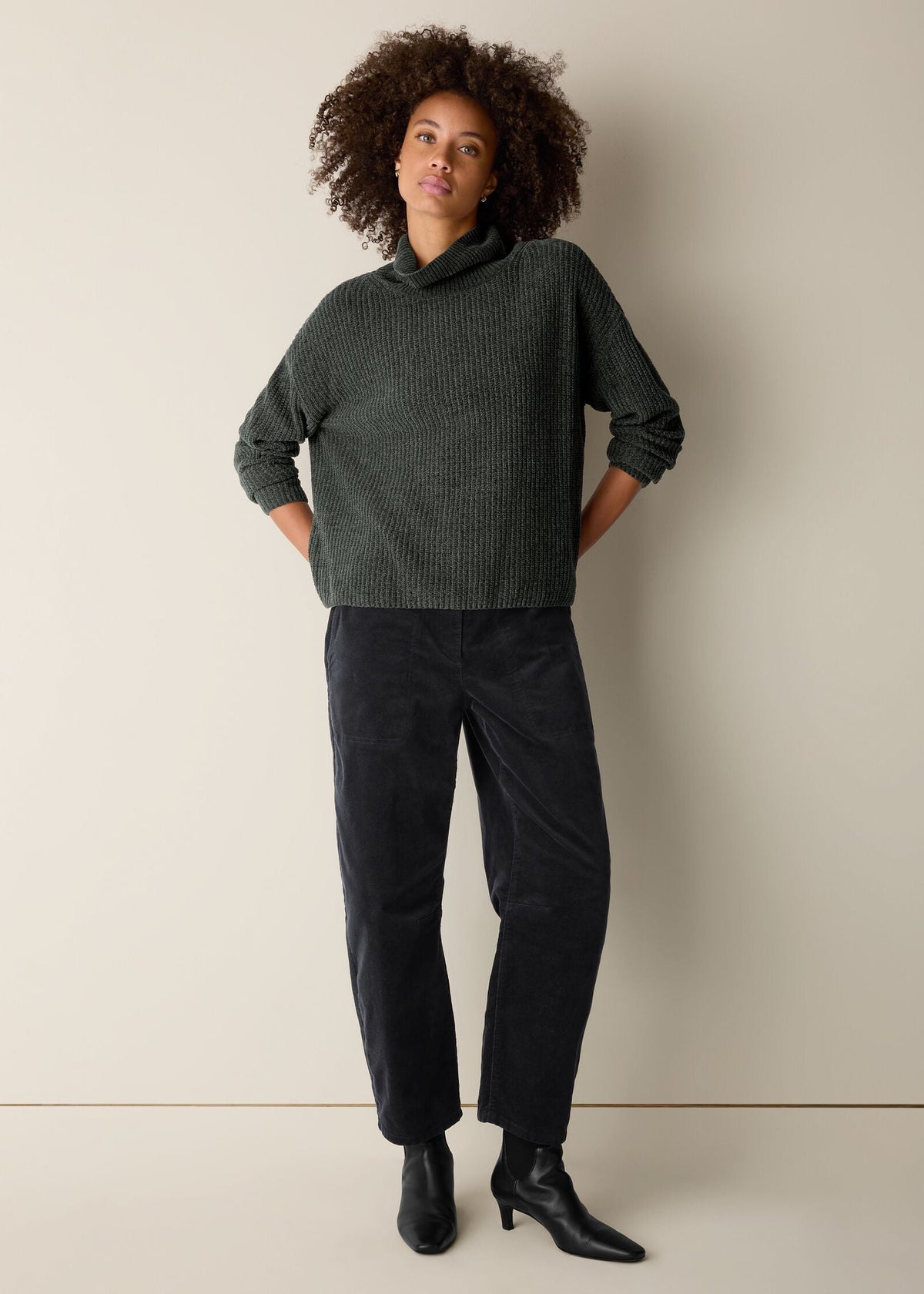 A woman stands against a plain beige studio wall wearing a dark green ribbed cowl-neck sweater, dark charcoal corduroy pants, and black ankle boots.