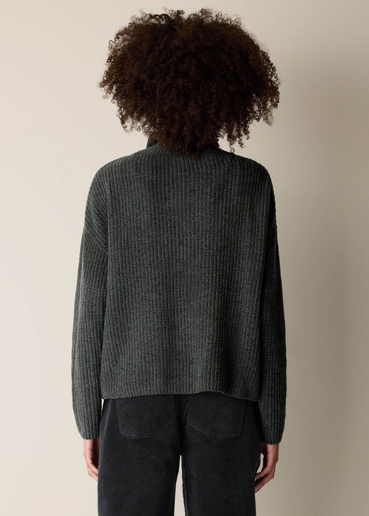 A person shown from the back wearing a dark olive green ribbed knit sweater and dark pants against a plain beige studio background.