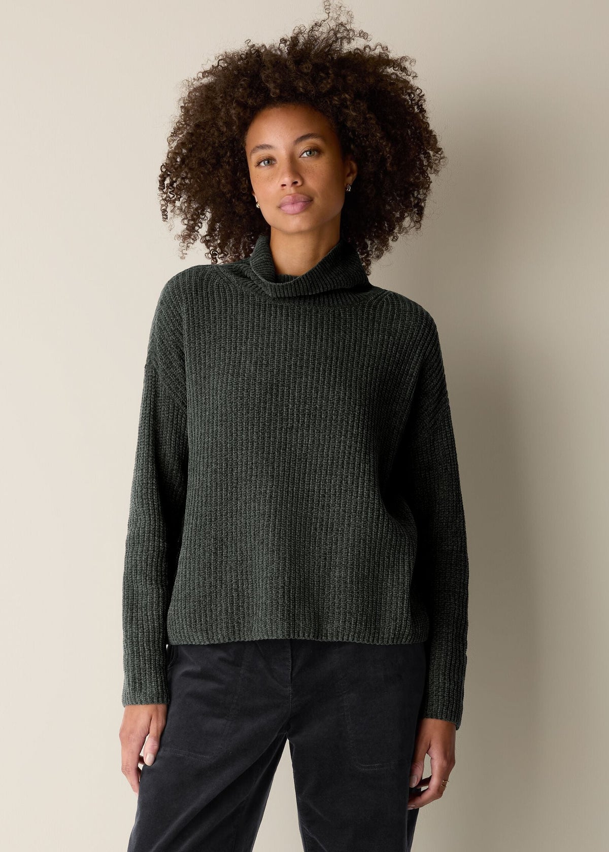 A woman with curly dark hair wearing a dark green ribbed turtleneck sweater and black pants, standing against a plain beige wall.