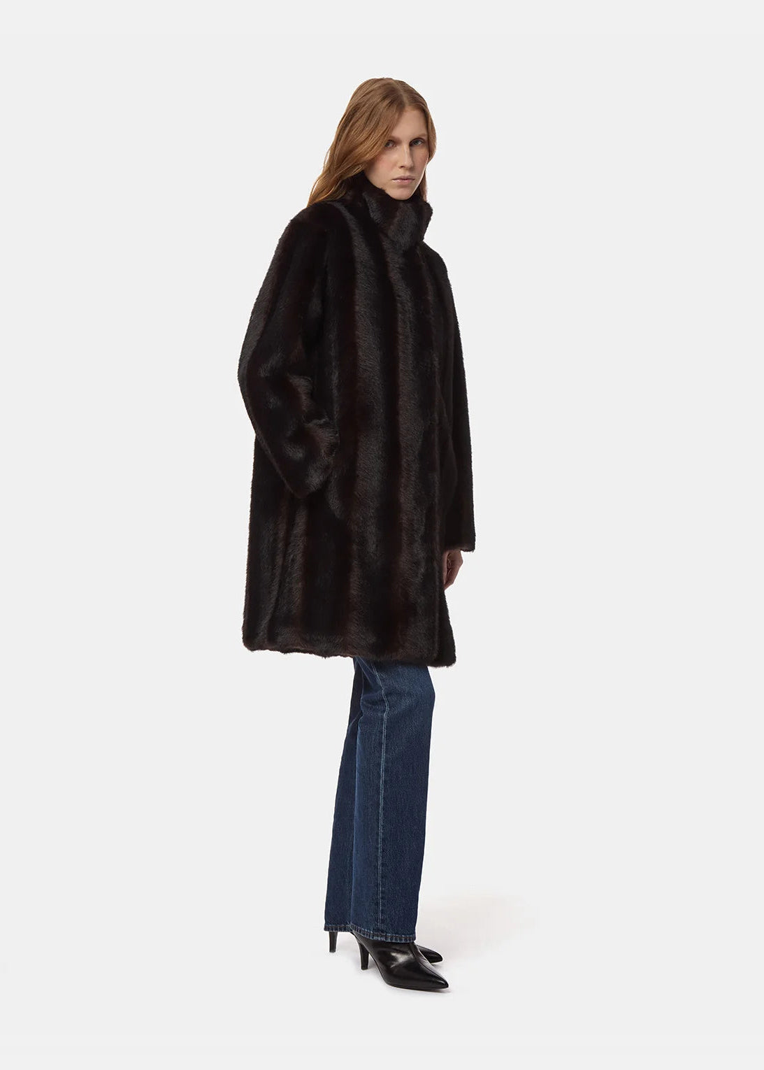A model wearing a long black faux fur coat with a high collar, paired with blue jeans and black pointed-toe heels, photographed against a solid light gray background.