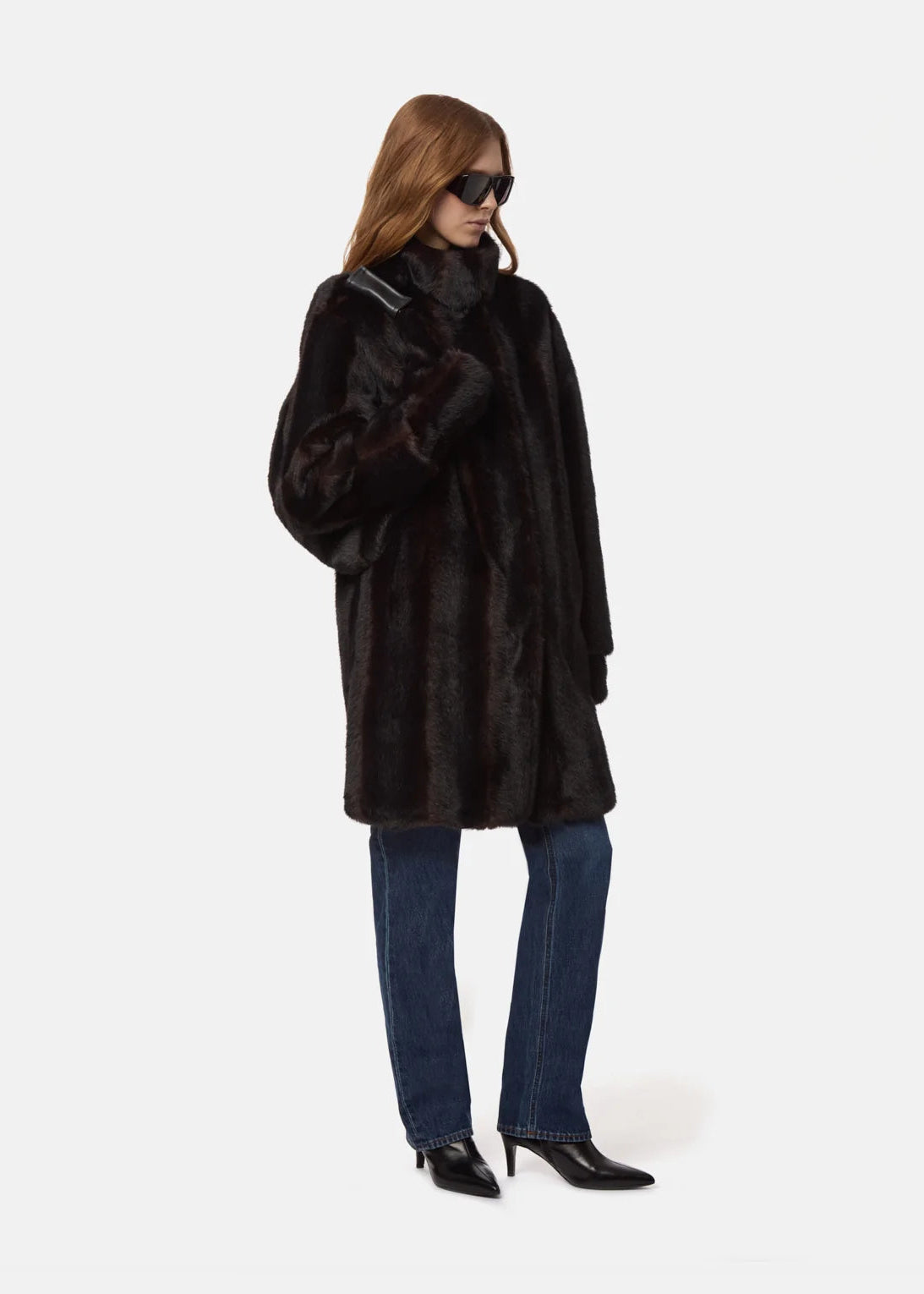 A model wearing a long black faux fur coat, blue jeans, and black pointed-toe heels, photographed against a solid light gray background.