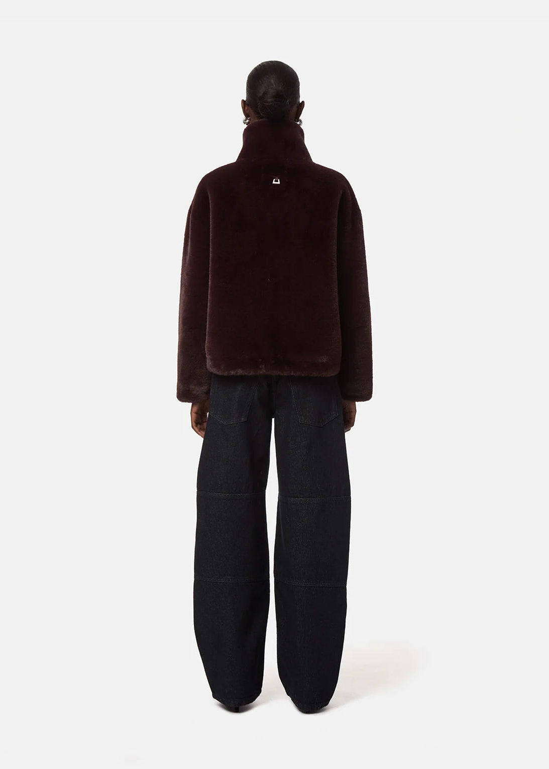 A model shown from the back wearing a dark burgundy faux-fur jacket and wide-leg dark trousers, photographed against a clean white studio background.