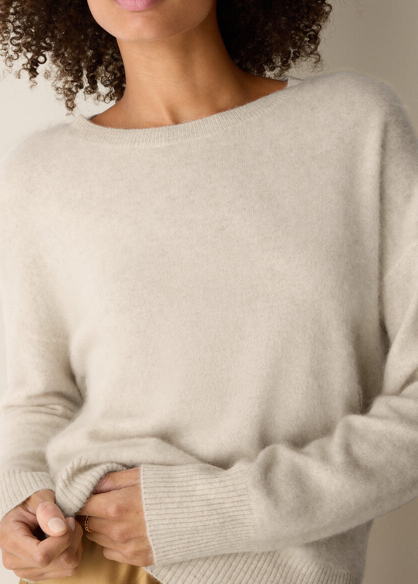 A close-up of a person wearing a soft, light beige knit sweater with a wide boat neckline and ribbed cuffs, shown against a plain light background.