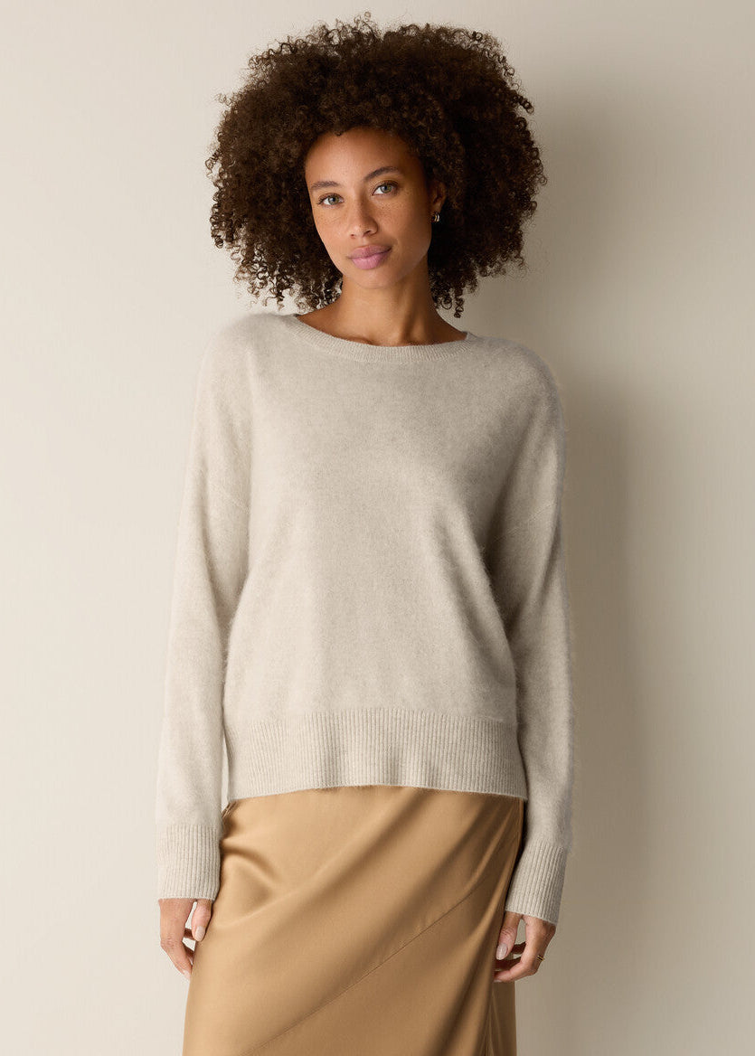 A woman with curly dark hair wearing a light beige knit sweater and a tan satin skirt, standing against a plain beige wall.