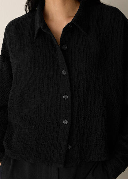 A close-up of a person wearing a black textured button-front collared top or cardigan with long sleeves, shown against a plain light background.