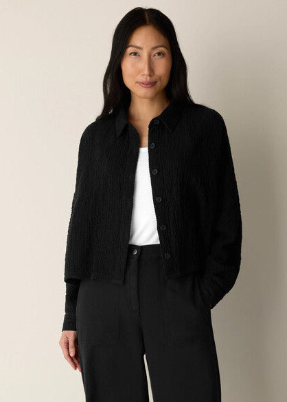 A woman modeling a black textured button-front cardigan over a white top and black trousers, standing against a plain light beige studio background.