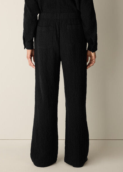 A person shown from the back wearing a black textured long-sleeve top and matching black wide-leg trousers against a plain light beige studio background.