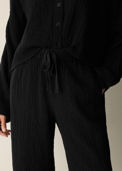 A close-up of a person wearing a black textured crinkle knit matching set, featuring a long-sleeve button-front top and wide-leg pants with a drawstring waist.