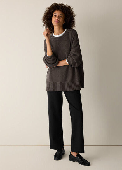 A woman modeling a dark brown oversized knit sweater with a white crew neck trim, paired with black wide-leg trousers and black leather loafers, against a plain beige studio background.
