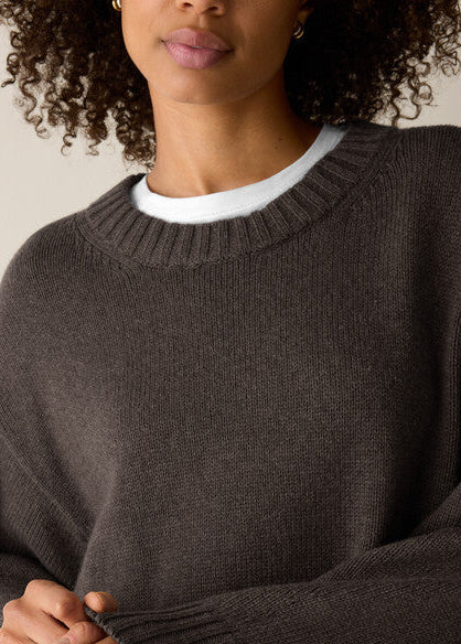 A close-up of a person wearing a dark brown ribbed knit sweater over a white crew-neck undershirt, with curly hair and a neutral beige background.