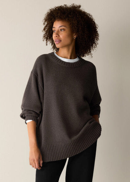 A woman with curly dark hair wearing an oversized dark brown knit sweater over a white undershirt and black pants, standing against a plain light beige studio background.