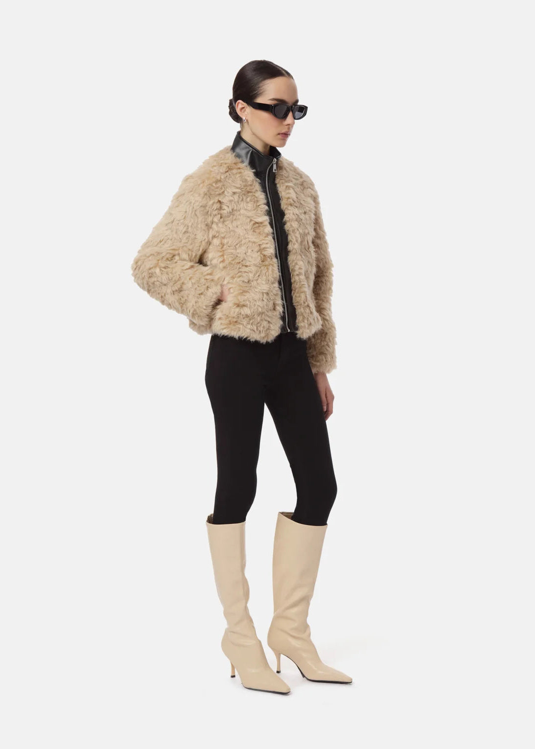 A model wearing a cropped beige shaggy faux-fur jacket with a black collar and zipper, paired with black leggings and beige knee-high pointed-toe boots, on a light gray background.
