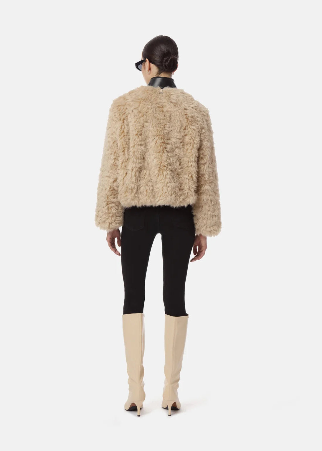 A model shown from the back wearing a beige shaggy cropped jacket, black skinny pants, and beige knee-high boots against a light gray studio background.
