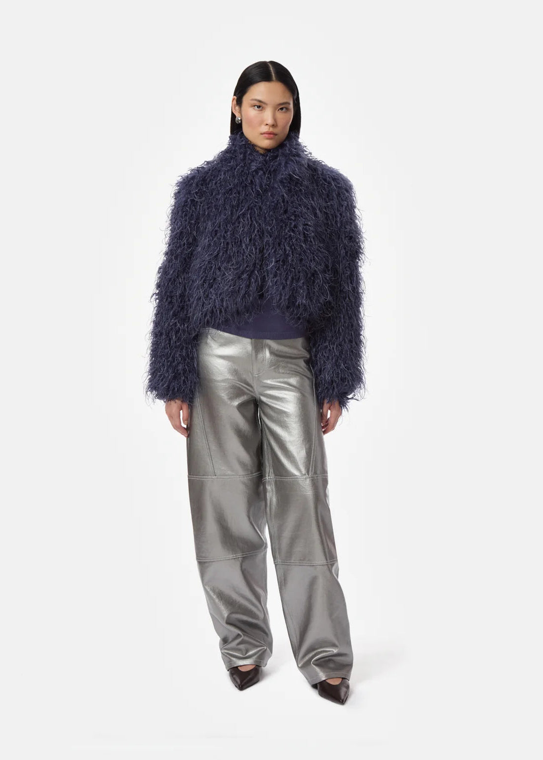 A model stands against a light gray studio background wearing a dark navy blue shaggy cropped jacket, a dark top, shiny metallic silver pants, and dark pointed shoes.