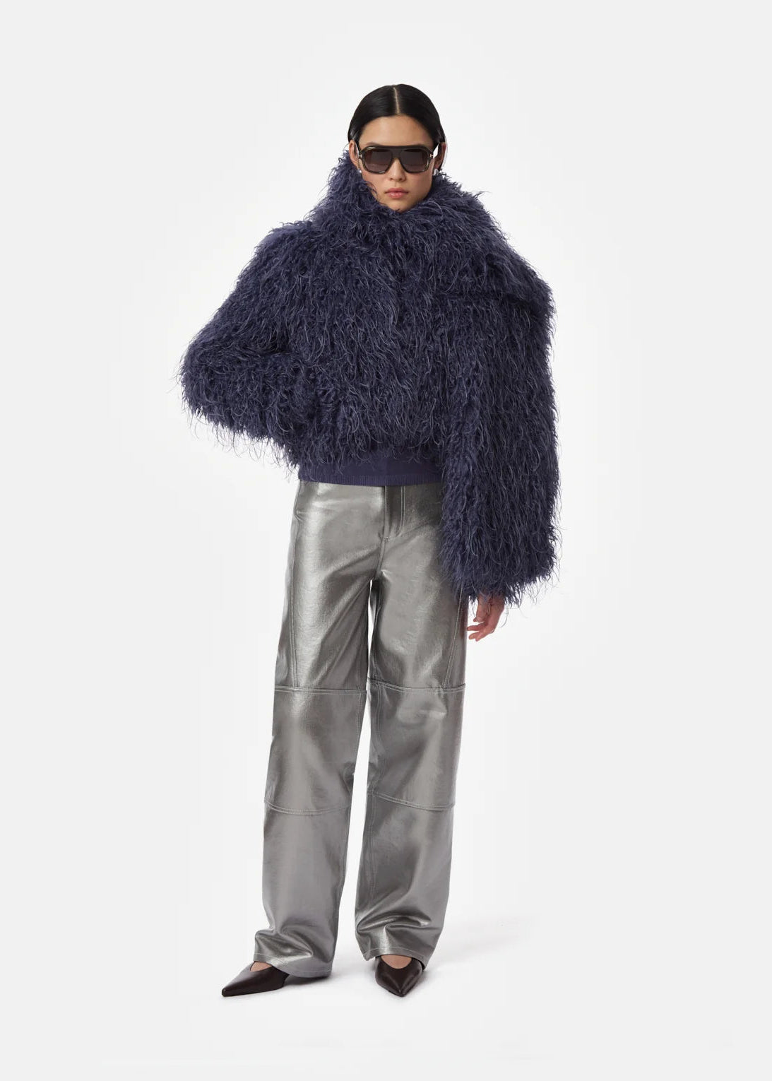 A model wearing a voluminous, dark navy blue shaggy faux-fur cropped jacket, metallic silver wide-leg trousers, and black pointed-toe flats, photographed against a seamless light gray studio background.
