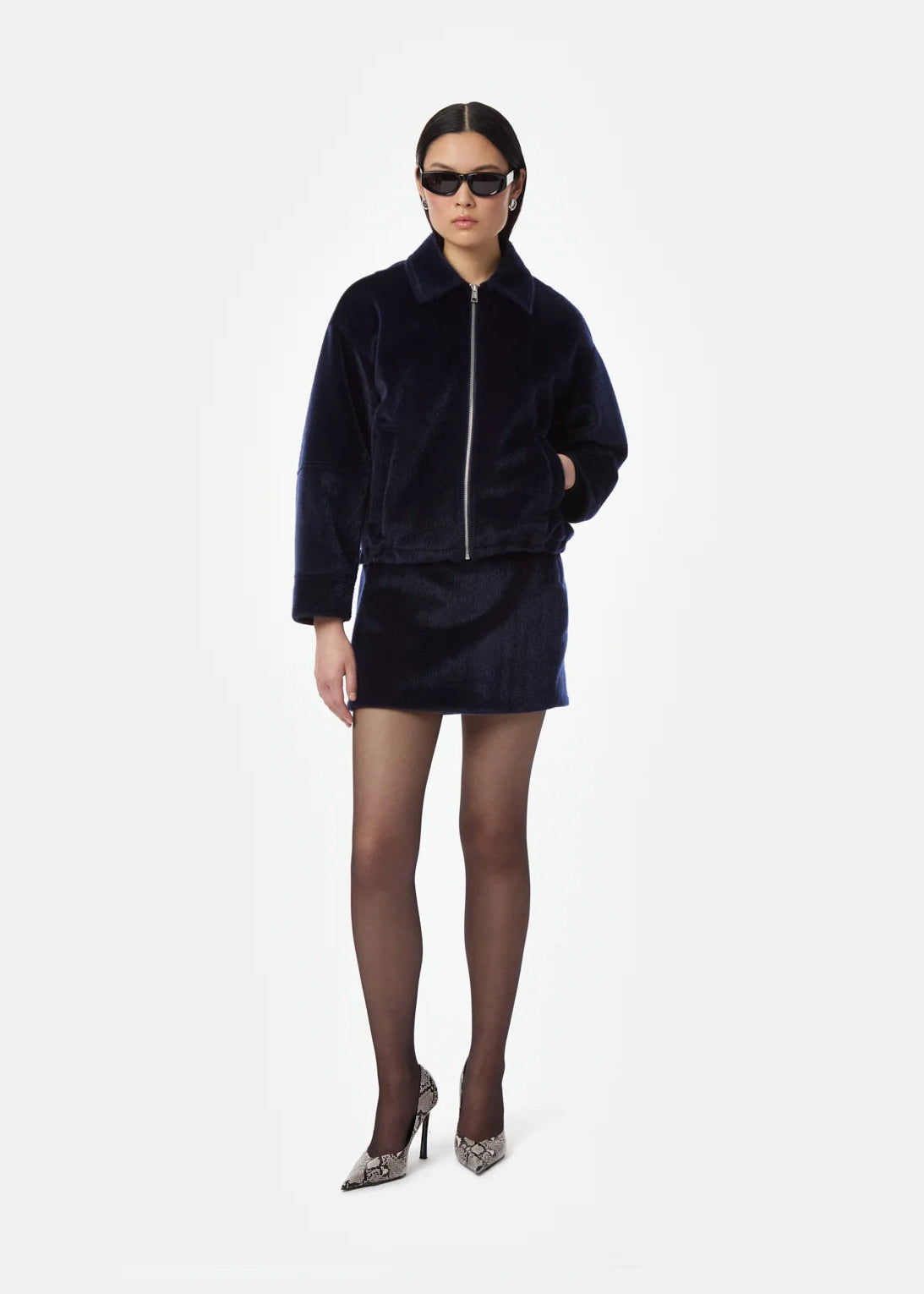 A model wearing a dark navy blue velvet jacket and matching mini skirt set, styled with black tights and snakeskin-patterned heels against a light gray background.