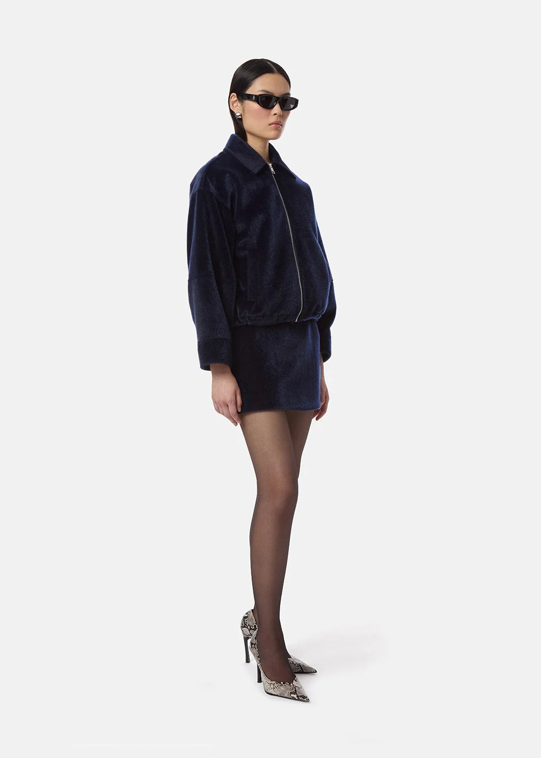 A model wearing a dark navy blue velvet jacket and matching mini skirt set, styled with black tights, patterned high heels, and sunglasses, against a plain light gray background.